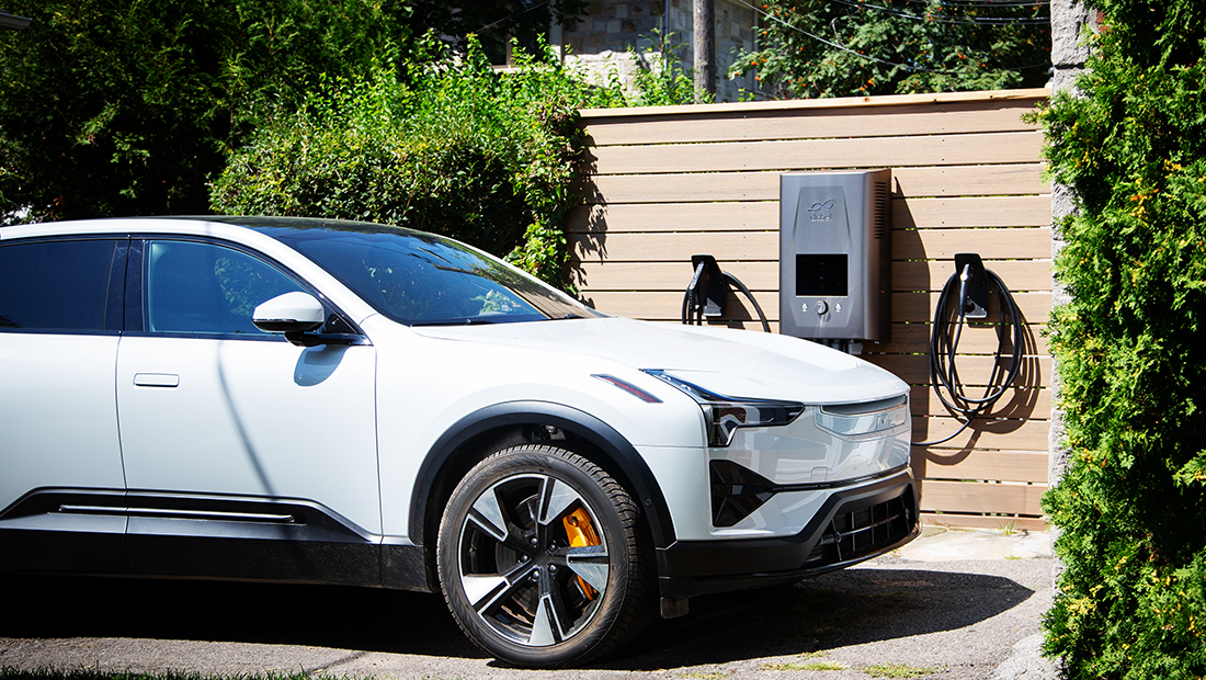 A white electric vehicle parked in a driveway, connected to a dcbel Ara Home Energy Station mounted on a wooden wall for charging, surrounded by greenery.