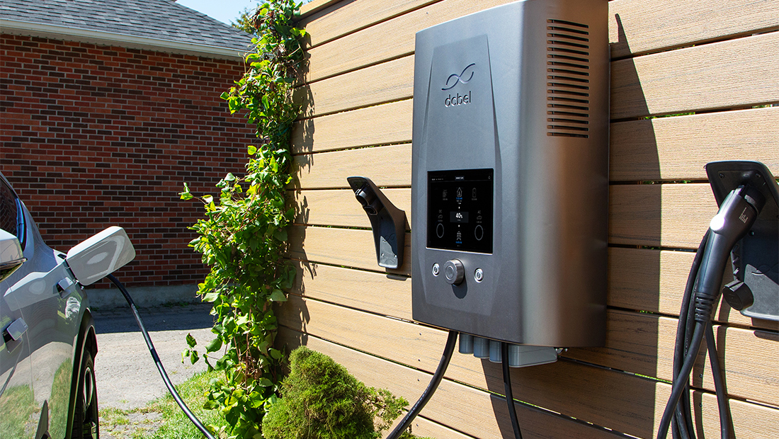 Wall-mounted dcbel Ara home energy system installed outdoors, connected to an electric vehicle charging cable beside a residential driveway.